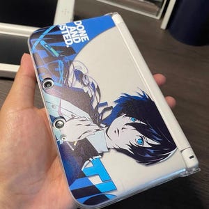 May include: A white and blue Nintendo 3DS handheld game console with a custom cover featuring anime artwork. The cover has a blue and white design with the text "DONE AND ISTED" and a character illustration.