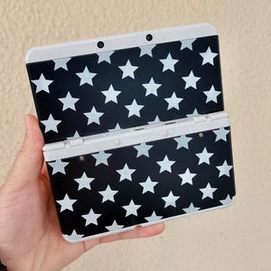 May include: A handheld games console featuring a black and white star design. The console is open, showing the screen and controls. The device is held in a hand.