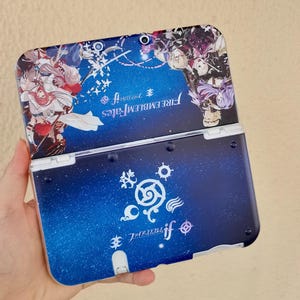Puede incluir: Funda azul y blanca para Nintendo 3DS con personajes y texto del juego "Fire Emblem Fates". La funda tiene un fondo estrellado y un diseño abatible.