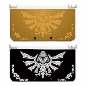 Puede incluir: Dos consolas Nintendo 3DS, una dorada y otra negra, cada una con un diseño de cresta estilizado. La consola dorada tiene una cresta dorada, mientras que la consola negra tiene una cresta blanca. Ambas consolas tienen una parte superior de plástico transparente.