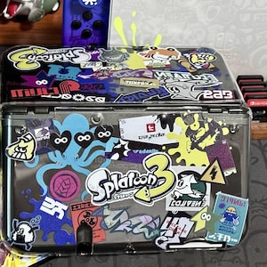 Könnte beinhalten: Eine transparente Kunststoffhülle mit bunten Aufklebern des Spiels "Splatoon 3", einschließlich Logo und Charakteren. Die Hülle enthält eine tragbare Spielkonsole und Spielkassetten mit Aufklebern in Blau, Gelb und anderen Farben.