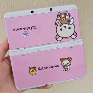 May include: Pink Nintendo 3DS handheld game console with white trim. The console features Rilakkuma character graphics and text. The top of the console is open, revealing the screen and buttons.