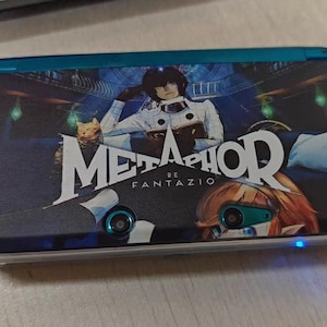 May include: A teal handheld gaming console featuring a custom skin with the Metaphor ReFantazio logo, a character in a white uniform, and an orange-haired figure. The device has two camera lenses on the front and a small glowing blue light indicator.