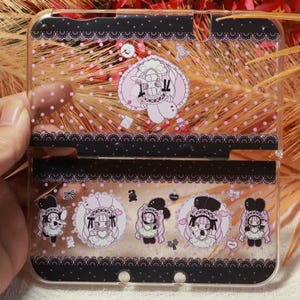 Puede incluir: Funda transparente para Nintendo 3DS con diseño de encaje negro y lunares rosas. La funda presenta ilustraciones de un personaje de dibujos animados con un atuendo rosa y blanco. La funda está abierta, revelando el interior.