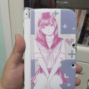 Puede incluir: Una consola de juegos portátil blanca con una funda protectora con una ilustración de estilo anime rosa y blanco de una chica con uniforme escolar. La funda tiene un fondo morado claro con signos más blancos.