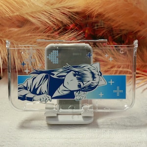 May include: Clear plastic phone stand with a blue and white anime-style illustration. The stand has a rectangular design with a blue stripe and white cross accents. A phone is placed inside the stand.