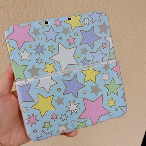 Op de afbeelding: Een handheld gameconsole met een lichtblauwe hoes met een patroon van kleurrijke sterren in roze, geel, groen, paars en wit. De console is open en toont het scherm en de knoppen.