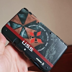 May include: Black and red handheld gaming console with the text "U.S.S." and "Umbrella Security Service Tactical Bag Limited". The console features a graphic design with a red cross and geometric shapes.