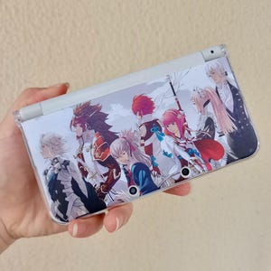 Peut inclure: Une console de jeu portable Nintendo 3DS blanche avec une coque personnalisée représentant un groupe de personnages de style anime. La coque a un fond clair avec plusieurs personnages en tenues variées. La console est tenue dans une main.