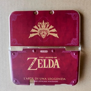 May include: A burgundy Nintendo 3DS case featuring gold lettering and graphics from "The Legend of Zelda." The case is open, revealing the interior. The text "L'ARTE DI UNA LEGGENDA" is also visible.