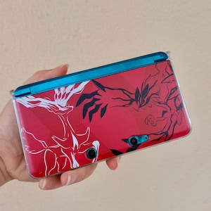 May include: A handheld gaming console with a red cover featuring white and black graphic designs. The console has a blue top and is held in a hand. The console has two camera lenses and a clear protective case.