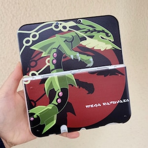 May include: A black and red Nintendo 3DS case featuring a green and black dragon design with the text "MEGA RAYQUAZA". The case is open, revealing the screen and buttons.