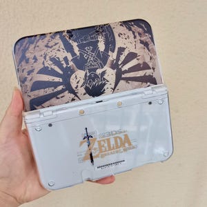 May include: A white Nintendo 3DS LL handheld gaming console with a decorative cover featuring a black and brown graphic design. The cover depicts a character from the Zelda series. The console's back has the text "ZELDA BREATH OF THE WILD".