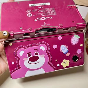 May include: A pink iQue DS handheld game console with a clear protective cover. The cover features a cartoon bear, strawberry, and other graphics. The console has a dual-screen design and various buttons.