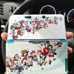 May include: A clear plastic case for a Nintendo 3DS with a colourful design featuring cartoon characters from the Pokemon video game series. The case has a white background with a blue border and a design of various Pokemon characters on the front and back.
