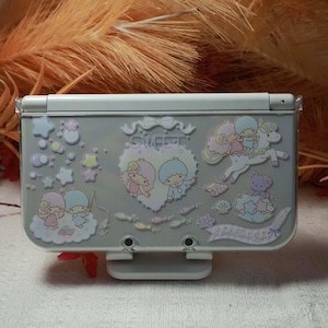 Puede incluir: Una funda gris claro para Nintendo 3DS con un diseño de dibujos animados colorido. El diseño incluye personajes de dibujos animados, estrellas y el texto "Sweet" y "Little Twin Stars". La funda tiene una cubierta de plástico transparente.