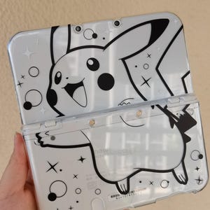 Op de afbeelding: Witte Nintendo 3DS-hoes met een zwart-wit Pikachu-ontwerp. De hoes is open en toont de binnenkant. Het ontwerp bevat Pikachu's gezicht en lichaam, plus sterren en andere decoratieve elementen.