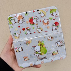 Könnte beinhalten: Hellgraue Nintendo 3DS-Handheld-Konsole mit einem Cartoon-Charakter-Design. Die obere und untere Hälfte der Konsole sind mit Bildern des Charakters, Früchten und Polka-Dots verziert. Die Konsole ist geöffnet und zeigt die Bildschirme und Tasten.