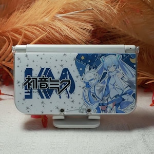 May include: White Nintendo 3DS console with a blue and white graphic featuring Japanese text and an anime character. The design includes snowflakes and stars. The console is resting on a white stand.