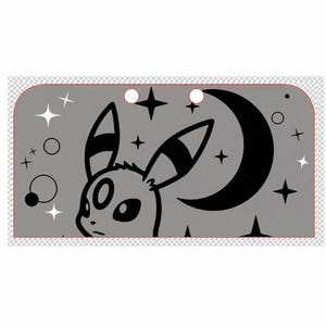 Puede incluir: Un accesorio rectangular gris con un diseño de personaje de dibujos animados en blanco y negro. El diseño incluye una luna creciente, estrellas y una cara de animal estilizada. El accesorio tiene dos recortes circulares en la parte superior.