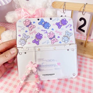 May include: A white Nintendo 3DS LL console with a clear case featuring a pink and purple teddy bear design. The case has the words "Cure", "Baby", and "Love" printed on it. The console is attached to a pink and white beaded charm.