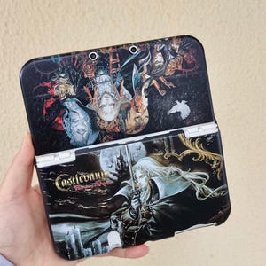 May include: A black Nintendo 3DS case featuring artwork from the Castlevania series. The case is open, revealing detailed illustrations of characters and scenes. The text "Castlevania: Harmony of Dissonance" is visible on the front. The case has a white hinge.