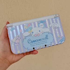 Puede incluir: Una funda para Nintendo 3DS a rayas azul claro y blanco con el personaje Cinnamoroll. La funda tiene un diseño de dibujos animados con una piruleta, estrellas y el texto "Cinnamoroll". La funda se sostiene en una mano.