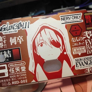 Könnte beinhalten: Eine braune und rote Nintendo 3DS-Hülle mit einer weißen und roten Anime-Charakter-Illustration. Die Hülle hat japanischen Text und die Worte "NEON GENESIS EVANGELION PLATINUM".