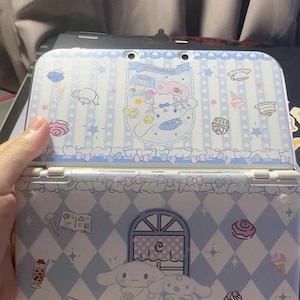 Peut inclure: Une coque de Nintendo 3DS bleu clair avec un design Cinnamoroll. Le boîtier présente un motif en losanges avec des illustrations de desserts et de personnages Cinnamoroll. La partie supérieure comporte une bannière avec du texte et des illustrations de personnages.
