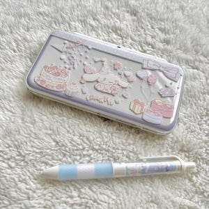 Puede incluir: Una funda blanca y lavanda con un diseño de personaje de dibujos animados y la palabra "PARTY". También hay un bolígrafo a juego con rayas azules y un diseño de dibujos animados. La funda presenta imágenes de un pastel, cupcakes y regalos.