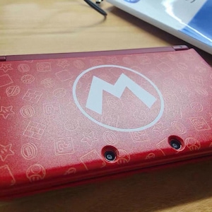 Puede incluir: Consola de juegos portátil Nintendo 3DS roja con un diseño de Mario. La consola tiene un exterior rojo con un patrón de iconos relacionados con Mario y un gran logotipo "M" blanco. El dispositivo está cerrado y sobre una superficie de madera.