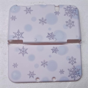 Puede incluir: Una funda 3DS de color lila claro con un diseño de copo de nieve. La funda tiene un fondo lila claro con copos de nieve dispersos y círculos azul claro. Los copos de nieve son de un tono púrpura más oscuro.