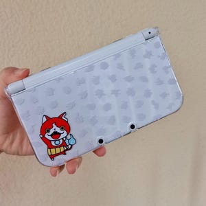 Op de afbeelding: Een lichtblauwe Nintendo 3DS handheld gameconsole met een doorzichtige beschermhoes. De console heeft een cartoon karakter sticker van een rood katachtig wezen met een blauwe vlam. De achterkant heeft een patroon van kleine, lichtgrijze vormen.