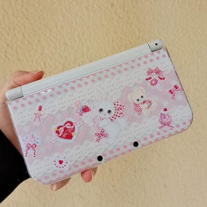 May include: A white and pink Nintendo 3DS case featuring a cute bunny, teddy bear, and heart design. The case has a polka dot pattern with lace accents and decorative bows. The overall theme is a sweet, kawaii aesthetic.