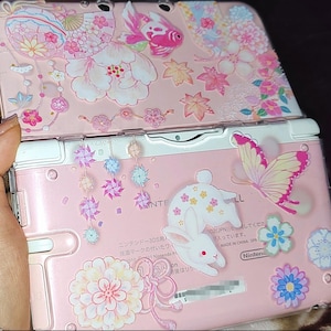 May include: A pink Nintendo 3DS handheld game console with a decorative floral and butterfly design. The top cover features a pink and white floral pattern, while the bottom has a white rabbit and butterfly sticker accents.