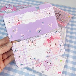 May include: A lilac and white Nintendo DS case with a Bonbonribbon design. The case features a gingham pattern, lace trim, and illustrations of bunnies with bows. The text "Bonbonribbon" and "Girly-cute cherry accessories" are also visible.