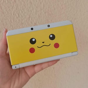 Puede incluir: Una consola de juegos portátil de color amarillo y gris claro con un gráfico de la cara de Pikachu en la cubierta frontal. El diseño incluye dos ojos negros, una nariz pequeña, una boca curva y dos mejillas rojas circulares.