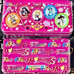 Può includere: Una custodia rosa per Nintendo DS con personaggi di Sailor Moon. La custodia è decorata con ritratti circolari di personaggi, stelle e testo giapponese. La custodia superiore ha una copertura in plastica trasparente.