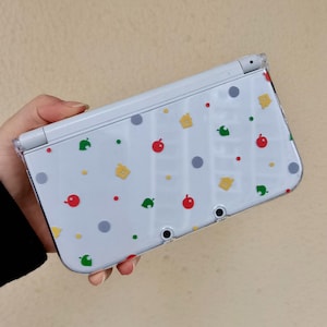 Może przedstawiać: Biała konsola do gier przenośna Nintendo 3DS z przezroczystą osłoną ochronną. Konsola jest ozdobiona wzorem czerwonych jabłek, zielonych liści, żółtych domów i szarych kropek.