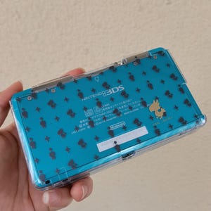 以下が含まれることがあります： 透明な保護ケースが付いたティールカラーのNintendo 3DS携帯ゲーム機。本体にはチョコボと星の小さな黒いシルエットが描かれており、背面に金色のチョコボのアイコンとFinal Fantasyの文字がプリントされています。