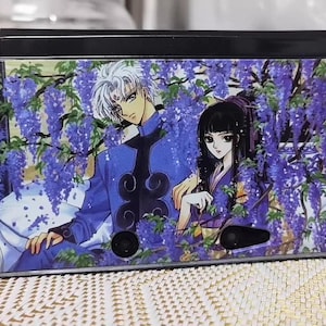 Peut inclure: Une console de jeu portable avec une illustration de style anime. Le design présente deux personnages au milieu de fleurs de glycine violettes. La console a un dessus noir et un devant transparent et brillant.