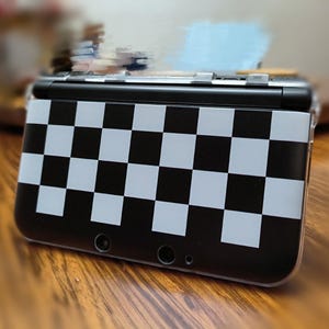 May include: A black and white checkered Nintendo 3DS handheld gaming console. The case features a classic checkerboard pattern, with alternating black and white squares. The console is on a wooden surface.