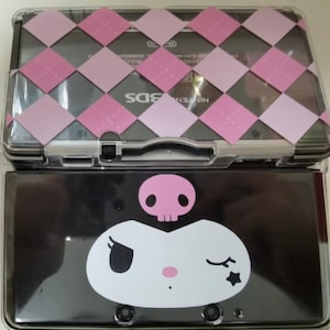 Op de afbeelding: Een zwarte Nintendo 3DS-console met een doorzichtige beschermhoes. De bovenste hoes heeft een roze en wit ruitpatroon. De onderste hoes heeft een wit cartoongezicht met een schedel en een ster.