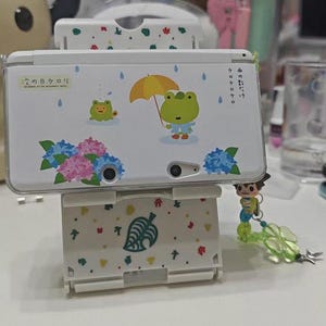 Könnte beinhalten: Eine weiße Nintendo 3DS-Konsole mit individuellem Design, das einen Frosch mit Regenschirm, blaue und rosa Blumen sowie japanischen Text zeigt. Die Konsole steht auf einem passenden Ständer mit Blattmuster.