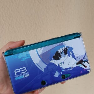 Könnte beinhalten: Blau-türkisfarbene Nintendo 3DS-Handheld-Konsole mit Artwork aus "Persona 3 Reload". Das Design zeigt eine Charakterillustration und das Logo des Spiels. Die Konsole wird in einer Hand gehalten.