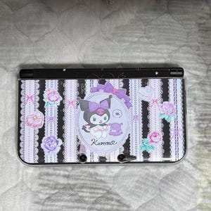 Puede incluir: Consola Nintendo 3DS en blanco y negro con diseño de Kuromi. El diseño incluye a Kuromi, lazos, corazones y detalles florales. La consola tiene una parte superior negra y está decorada con rayas verticales.