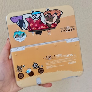Op de afbeelding: Een beige New Nintendo 3DS LL handheld spelcomputer met Monster Hunter Airou karakterafbeeldingen. De bovenkant toont katten die ontspannen onder een kotatsu, terwijl de onderkant kleine katillustraties en tekst bevat.