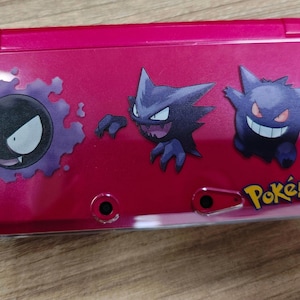 Puede incluir: Una consola de juegos portátil Nintendo 3DS magenta con tres personajes Pokémon: Gastly, Haunter y Gengar. La palabra "Pokémon" está impresa en amarillo. La consola tiene un acabado brillante.