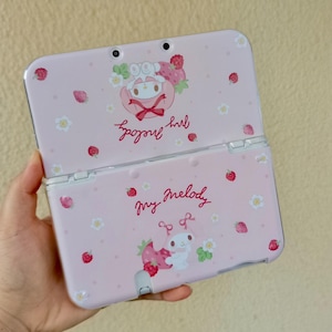May include: A pink Nintendo 3DS handheld game console with a My Melody design. The case features illustrations of My Melody, strawberries, and flowers. The words "My Melody" are written in cursive.