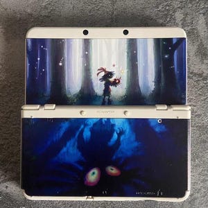 May include: A white Nintendo 3DS console with custom artwork. The top screen features a forest scene with a figure holding glowing orbs. The bottom screen displays a dark, blue-toned image with two glowing eyes. The console is open, revealing the screens.
